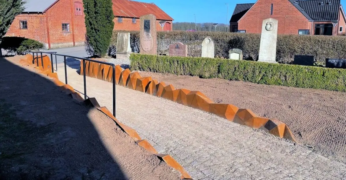 en usædvanlig stålkant af 452 forskellige trekant plade i 5 mm cortenstål, der er svejset som en stenvæg langs stien mod Elev Kirke