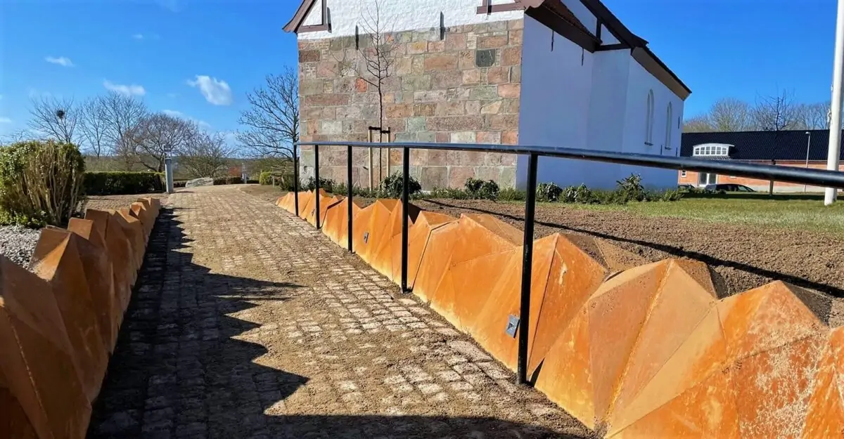 en usædvanlig stålkant af 452 forskellige trekant plade i 5 mm cortenstål, der er svejset som en stenvæg langs stien mod Elev Kirke
