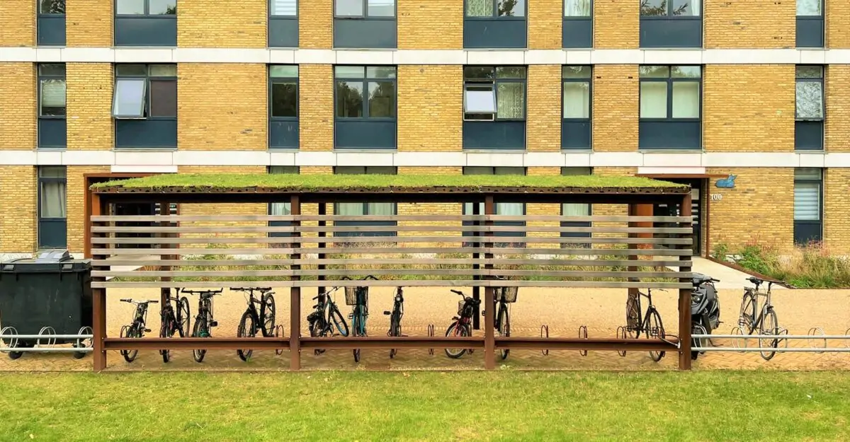 en modulopbygget cykeloverdækning i cortenstål med installeret cykelstativer og beklædning af mahogni planker, som står ved siden af en stor lejlighedsbygning i Vollsmose